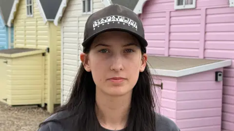 Jamie Niblock/BBC Leonie Powling stands on a stony beach, in front of multicoloured beach huts. She wears a dark-coloured jumper and black baseball cap.
