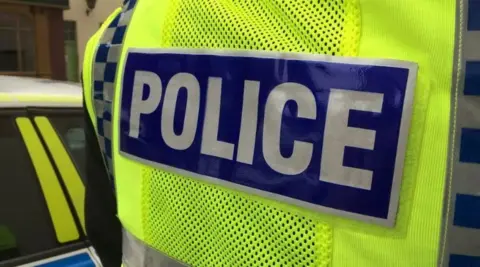 Close up picture of the back of a high vis police jacket as an officer stands next to a police car