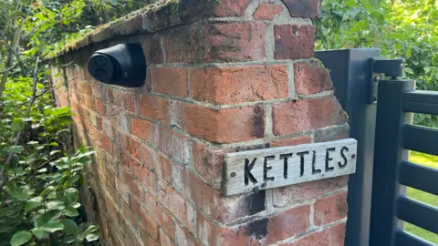 John Fairhall/BBC A brick wall with the sign 'Kettles' next to a brown wooden gate. On the other side is a big green bush