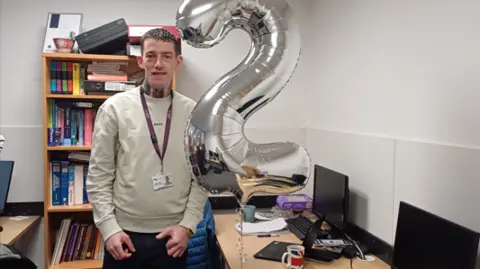 Mr Kelly is stood by a silver number two balloon marking his anniversary. He has a grey jumper on and jeans and is standing by a table in an office.