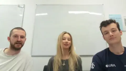 The team members are sitting in a room with a white board behind them. Toby has closely cropped hair, a neatly trimmed beard and he is wearing a white T-shirt. Manon has long blonde hair and she is wearing a dark green coloured top. Seb has dark hair and he is wearing a dark blue T-shirt.