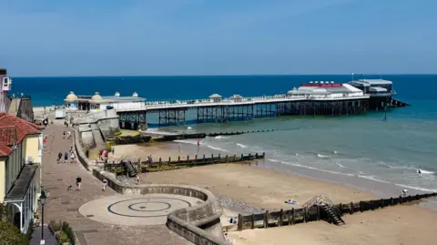 View from the promenade of Cromer beach looking out towards the pier. The pier stretches across the water which is light blue. People are walking along the promenade. 
