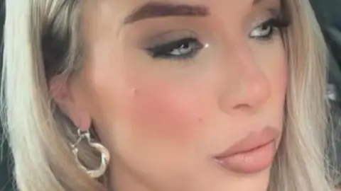 A close up image showing a young woman with blonde hair and gold earrings looking off camera