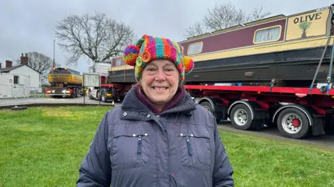 Diane Taylor, a woman in a multicoloured hat and blue coat is smiling at the camera and standing next to a haulage truck which carries a boat with the word Olive on it
