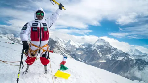 PA Media A man in a red and white snow suit, with two above knee prostheses stands atop Mount Everest holding his hand in the air to celebrate. 