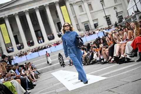 AFP via Getty Images A woman dressed in light blue denim walks down a catwalk in Trafalgar Square, London.