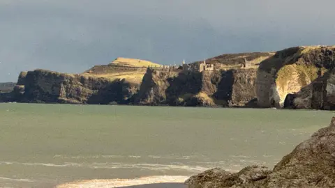 The ruins of Dunluce castle can be seen from afar, perched on top of a cliff. The ocean sits at the foot of the cliff, which stretches from the cliff to the camera. 