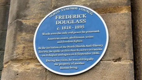 A blue circular commemorative plaque mounted on a stone church wall. The plaque, issued by North Tyneside Council, honours Frederick Douglass, describing him as an American orator, abolitionist, writer, and freedom fighter. It notes that Douglass spoke at the church twice in 1846 at the invitation of the North Shields Anti-Slavery Society and states that, during those visits, he was still legally enslaved.