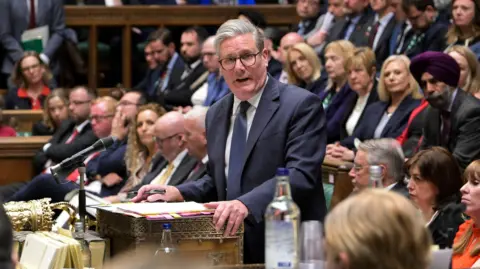 Keir Starmer stood in the House of Commons wearing a suit and tie. He is stood at the despatch box and is speaking with a folder in front of him. He is surrounded by members of his party who are sat to his sides and behind him.
