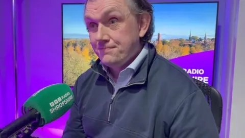 A man with blue shirt and top sat in a radio studio with purple background and a green microphone cover