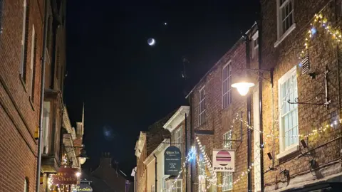 Gryning/Weather Watchers The moon and Venus shine down on a street in York. Stings of fairy lights are threaded alongthe buildings. Signs for Pure Hair and Quacks shop are visible. 