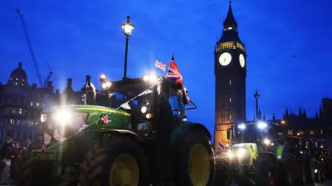 A tractor flying a union jack flag drives past Big Ben at night