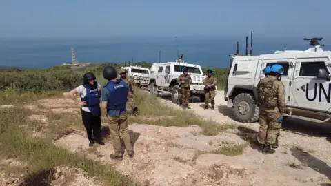 Seven people wearing helmets and bulletproof vests standing next to three white trucks with the words UN marked on the side