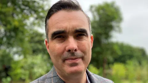 Martin Lovell, managing director of the Story of Emily, smiling at the camera in a grey blazer with trees in the background. 