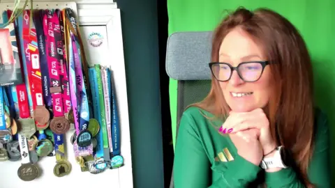 Sarah wearing glasses, a white sports watch and green running shirt. She is sitting next to her marathon medals. She is wearing black-rimmed glasses, has long auburn hair and is wearing pink nail varnish.