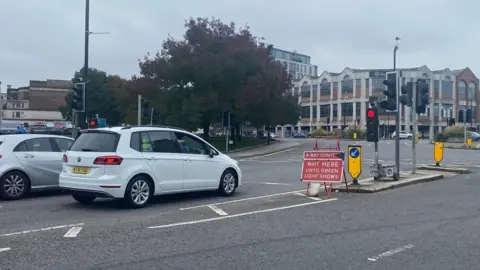 A white car waits at a junction where there is a red temporary traffic sign which reads: "Wait here until green light shows." Next to it is a temporary traffic light which is on red. Behind that are several permanent traffic lights which appear to be off