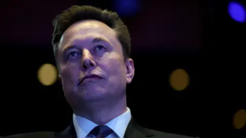 Reuters Elon Musk in a black suit and grey neck tie staring out with a bemused look on his face.