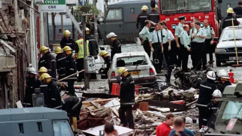 PA Media several emergency respondents stand on a road after a bomb. Derements and debris cover the road and road. Cars are damaged with cars outside the glass