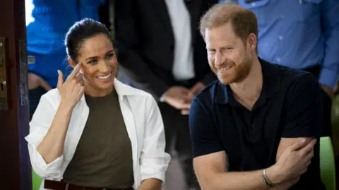 The Duke and Duchess of Sussex siting next to each other smiling at something.