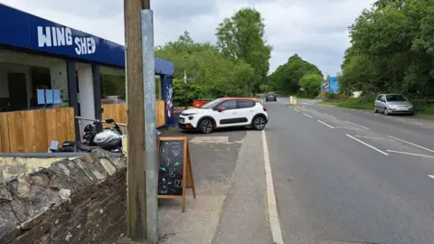 Alison Taylor A white car with a red roof parked outside Wing Shed in London Apprentice. It is blocking the pavement and has part of its rear end edging into the road. A blackboard sign saying 'open' is on the roadside.