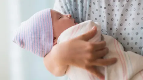 Getty Images A mother in a hospital gown holding a baby in her arms, which is covered in a blanket