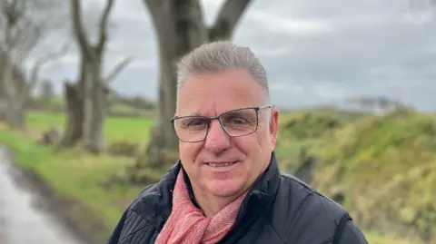 Graham Thompson has short grey hair swept back from his head and glasses. He is wearing a black coat and red scarf and is standing in front of one of the Dark Hedges trees 