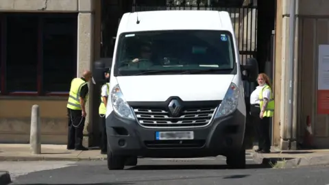 A white van drives away from a prison as security guards in high-visibility vests stand either side of it.