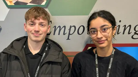 A teenage boy with wavy blond hair and a black coat beside a teenage girl with black hair tied back behind her head and large glasses and a black top