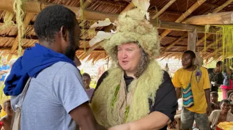 A person wearing a large ceremonial outfit made from natural fibres is standing inside a traditional thatched structure and shaking hands with another person. Several people are visible in the background, along with wooden posts, woven roofing, and hanging plant materials that suggest a community gathering or cultural event.