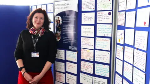 University of Bath Prof Turi King stands in front of the wall of plaques dedicated to the women at the university. She has short reddish-brown hair, a black top, a red skirt and is smiling at the camera.