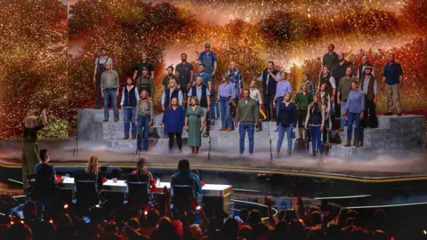 Will Rogers A large group of performers stands on tiered platforms on a brightly lit stage, appearing to sing together as a choir. A conductor stands at the front, guiding the performance. In front of the stage, a panel of judges sits at a table, watching. The background features warm, glowing lights and foliage-like scenery, while an audience in the foreground raises hands and watches the show.