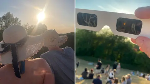 BBC/ Hannah Griffiths On the left, a man and a woman are facing away from the camera and observing the sun. Both are wearing straw-type hats. On the left is the view through a pair of eclipse glasses, with the Sun an orange dot on the right-hand lens. Beneath the glasses, families sit on the grass in groups watching the event