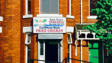 Family handout The Sam Sams Takeaway building is a red brick house with a large white sign hung above the door. There are white window frames on the building, and a red car is parked out at the front.
