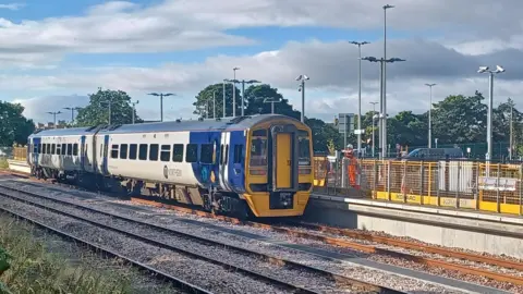 A blue and white, two-carriage Northern train pulled up to the station platform. There are yellow barriers erected on the platform, with a worker wearing a high-vis uniform standing on it too. There are about a dozen poles rising from the station grounds, with CCTV and street lights.