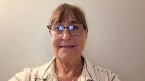 ANN SAVELL A head and shoulders picture of Ann Savell who is smiling and looking at the camera. She is just in front of a white wall. There is a slight shadow to the right of the picture.