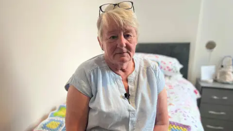 Woman with short blonde hair black glasses on head light blue blouse sat on single bed with blanket on top