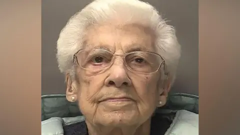 A mugshot of a 95-year-old woman with white hair and glasses, who looks at the camera with a straight face. She wears pearl earrings and a blue puffer-coat over a jumper. She also has hearing aids.