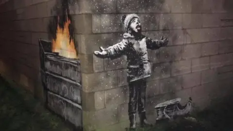 Scott Bamsey Banksy drawing across two walls at right angles - the right side shows a young child with his arms splayed, a sled at his feet and tongue out looking at what looks like falling snow. The left side reveals the snow is in fact ash from a fire burning in a dump truck.