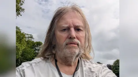 A selfie of John Adams. He has shoulder length light grey hair, a beard and bushy eyebrows. He is standing outdoors on a cloudy day beside a tree.