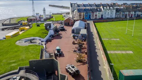 Heugh Battery Museum An aerial view of the museum. There are tanks and guns, pointing at the sea, to the left.
At the back there is a lighthouse, pier and a row of colourful houses. 