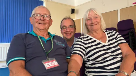 Graham has short grey hair and glasses, he is wearing a green and navy polo shirt, and a green hidden disability lanyard. Rick has short dark hair and glasses. Lorraine has short blonde hair and is wearing a black and white striped top.