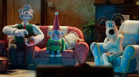 Wallace is sitting next to Norbot, a robotic gnome, on a red and white patterned sofa. Wallace is pointing a remote control at the TV. Gromit is sitting on a chair which is light blue with yellow dots. A piano, a pot plant and floral patterned wallpaper can be seen on the wall behind them.