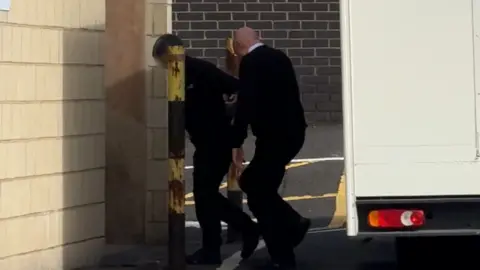A man being led away from a van by another man wearing a black jumper and a shirt. He is walking into a brick building.
