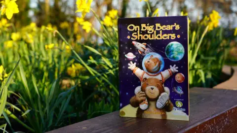 Thea Lawson The image shows a children's book in front of some daffodils. The book is called On Big Bears Shoulders, the cover is a space scene with two animated bears, a big bear with a smaller bear on its shoulders. The smaller bear is wearing a space suit and has its head in space. 