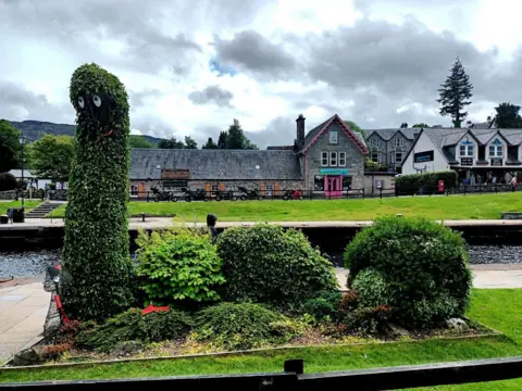Lynn Martin A group of bushes trimmed into the shape of the Loch Ness Monster. There are buildings and a park in the background.