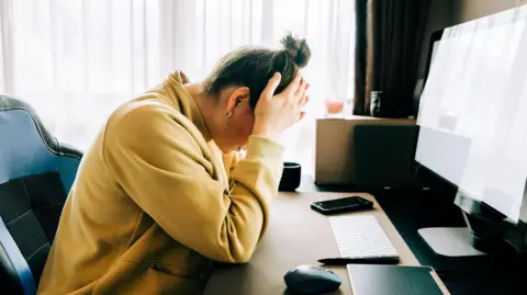 A woman wearing a yellow jacket and with her hair tied up sits at a desk with a keyboard and large monitor. The woman has her head in her hands.