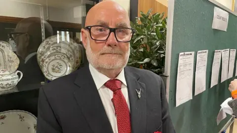 Steve Thornley - a bald man with glasses, a white beard, black jacket, white shirt and red tie - stands in front of a glass cabinet, which has crockery stored inside. 