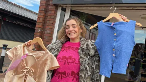 Fashion designer Victoria Ford with long brown wavy hair wearing large silver hooped earrings and a faux fur leopard skin coat and hot pink knitted jumper with dark red bows on it holds two of her designs up outside a charity shop. In her right hand (pictured left) is a gold ladies top on a coat hanger and in her left hand she holds up a blue sleeveless top on a coat hanger. She is smiling. 