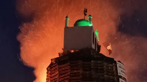 Handout A bonfire is on fire. An effigy of a model that looks like a mosque is on top with banners. It's night.