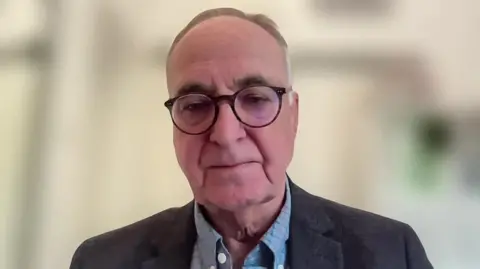 Professor Ted Cantle, who is wearing a grey blazer, blue shirt and glasses, is starring directly at the camera during an interview. The background has been blurred out.